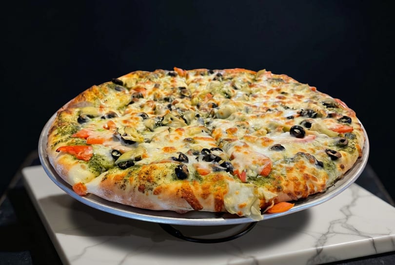 A whole pizza with pesto, cheese, tomatoes, black olives, and artichoke hearts on a metal pan.