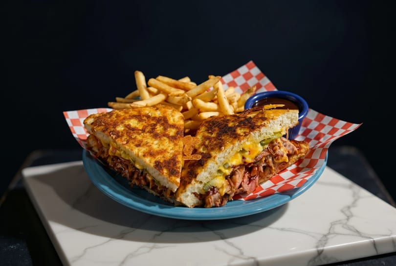 A grilled cheese sandwich with pulled pork and pickles, served with fries and BBQ sauce.