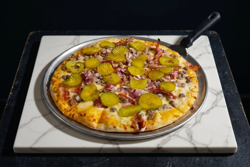 A whole pizza with pickles, ground meat, bacon, and red onions on a marble slab.